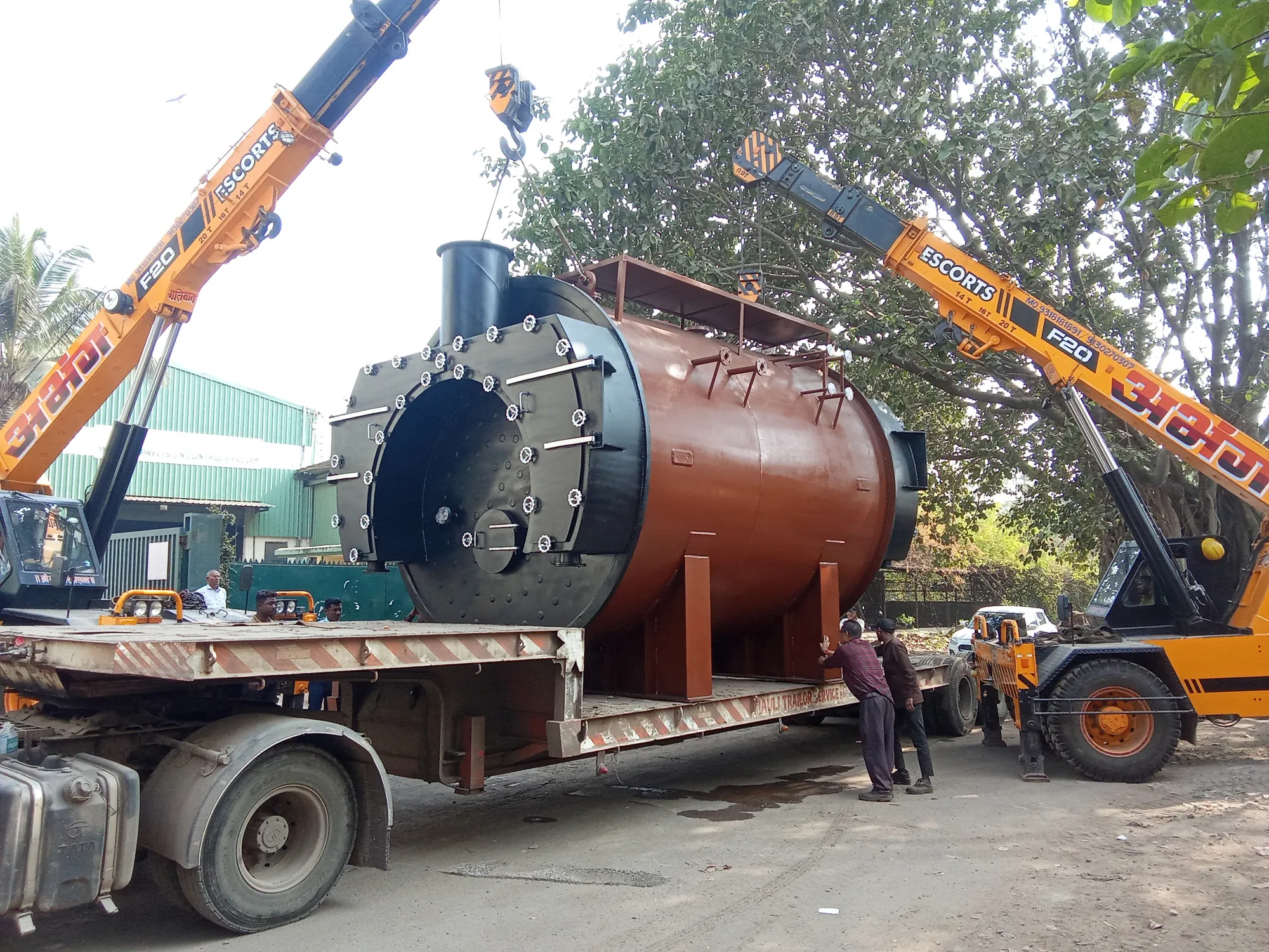 Two yellow cranes lift a large rust-red industrial boiler onto a low-bed trailer while the crew guides it into place.